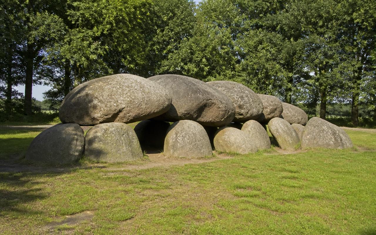 Hunebed van grote zwerfkeien op een grasveld aan de rand van een eikenbos in Drenthe in laat middaglicht.