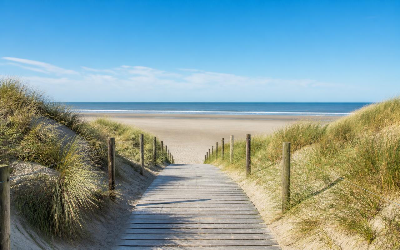 Houten vlonderpad door duinen met helmgras naar het brede Noordzeestrand bij Bredene.
