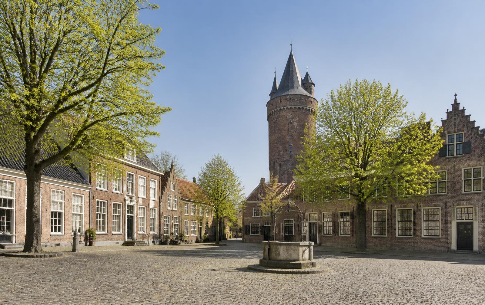 Geplaveid plein in 's-Heerenberg met bakstenen huizen en de ronde toren van Kasteel Huis Bergh achter de bomen.