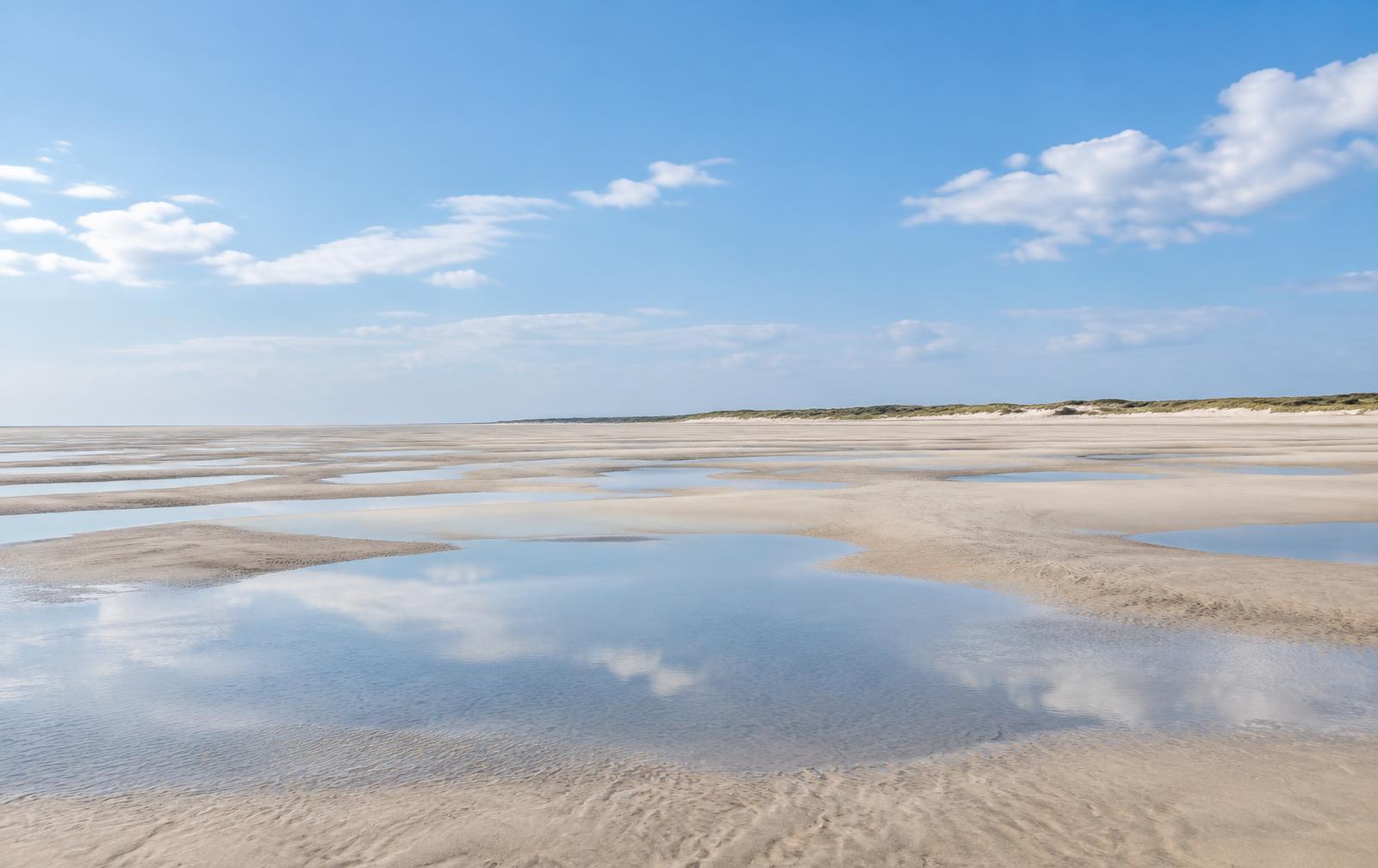 Uitgestrekte bleke zandvlakte met ondiepe plassen die de blauwe lucht en wolken weerspiegelen, met een lage duinrij in de verte.