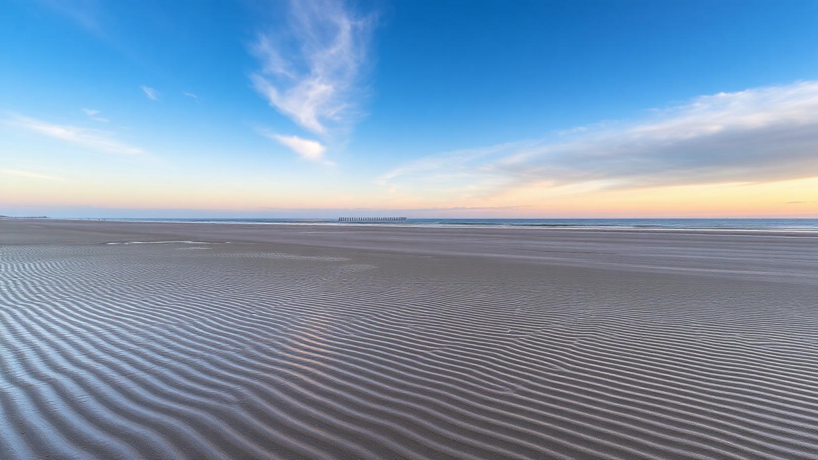 Breed Noordzeestrand bij eb met ribbels in het zand en golfbrekers in de verte onder een avondlucht.